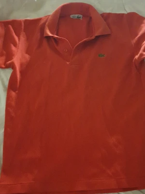 Röd pikétröja från Lacoste - Klassisk röd pikétröja från Lacoste med korta ärmar och den ikoniska gröna krokodilloggan på bröstet. Tröjan har krage och knappar vid halsen. Perfekt för en avslappnad och stilren look. Riktigt nice färg o material, storlek S,M typ