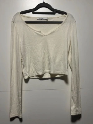 Vit croppad långärmad topp NA-KD - Säljer en vit croppad långärmad topp från NA-KD med v-ringning. Toppen har en enkel och stilren design, perfekt att matcha med höga jeans eller kjol. Materialet känns mjukt och lätt mot huden.