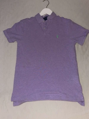 Oanvänd Lila pikétröja från Ralph Lauren - Säljer en oanvänd klassisk lila pikétröja från Ralph Lauren med korta ärmar och krage. Tröjan har två knappar vid halsen och den ikoniska gröna loggan broderad på bröstet. Perfekt till jeans eller shorts för en avslappnad men snygg stil. Nypris är på över 1500kr 