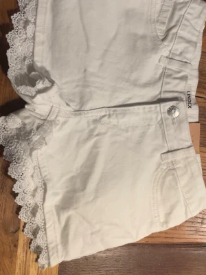 Vita spetsdetaljerade shorts Lindex - Säljer ett par vita shorts från Lindex med fina spetsdetaljer längs bensluten. De har normal passform, klassisk femficksdesign och knappstängning framtill. Perfekta för varma dagar och ger en söt touch till din outfit.