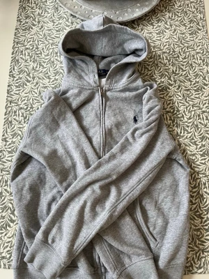 Grå zip-hoodie från Polo Ralph Lauren - Säljer en grå hoodie med dragkedja från Polo Ralph Lauren. Klassisk design med huva, två fickor fram och broderad logga på bröstet. Perfekt för en avslappnad och stilren look. Materialet är mjukt och bekvämt.