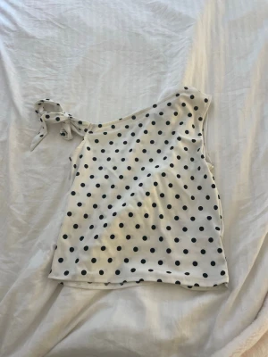 Prickig oneshoulder topp med knyt - Säljer en vit oneshoulder topp med svarta prickar och knyt på axeln. Toppen har en avslappnad passform och är perfekt för dig som gillar en lekfull och trendig stil. Supersnygg till både jeans och kjol!