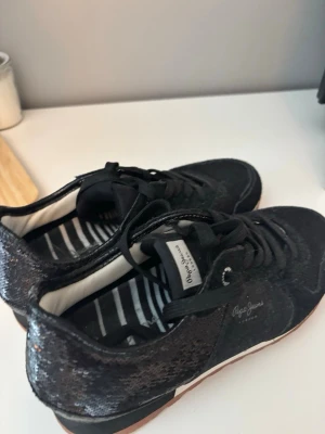 Svarta sneakers från Pepe Jeans - Snygga svarta sneakers från Pepe Jeans med glittrande paljettdetaljer och vita ränder på innersulan. Skorna har klassisk låg design, svarta skosnören och vit sula med brun kant. Perfekta för dig som vill sticka ut lite extra.
