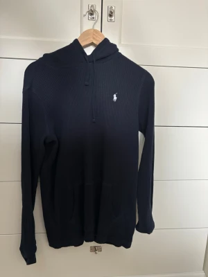 Mörkblå hoodie från Polo Ralph Lauren - Snygg mörkblå hoodie från Polo Ralph Lauren med broderad vit logga på bröstet. Tröjan har huva med dragsko, känguruficka och är tillverkad i ett ribbat, mjukt material. Perfekt för en avslappnad och stilren look.