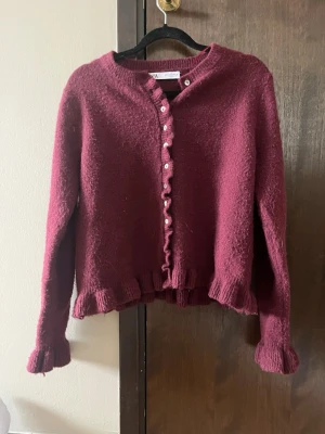 Vinröd stickad kofta med volangkant - Söt vinröd stickad kofta med volangkant och små vita knappar framtill. Koftan har långa ärmar och ribbade muddar vid ärmslut och nederkant. Materialet ser mjukt och fluffigt ut. Från Zara. Säljer för att den inte används. Storlek M.