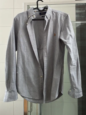 Grå skjorta från Polo Ralph Lauren - Snygg grå skjorta från Polo Ralph Lauren i slim fit-modell. Skjortan har klassisk krage med knappar, långa ärmar och det ikoniska broderade logotypen på bröstet. Perfekt för en stilren look.