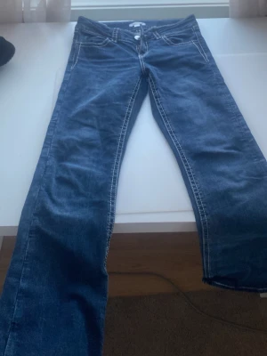 Blå bootcut jeans med broderi - Säljer ett par blå bootcut jeans med vita kontrastsömmar och snyggt broderade bakfickor med silverdetaljer. Jeansen har låg midja och klassisk femficksdesign. Perfekta för dig som gillar en avslappnad men ändå trendig look.