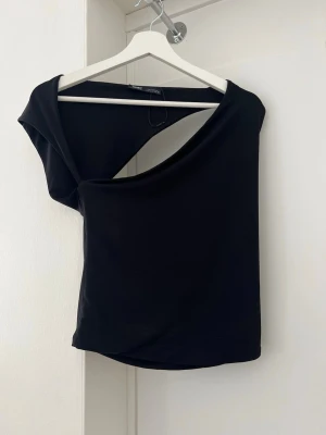 Svart oneshoulder topp från Gina Tricot - Trendig svart topp från Gina Tricot med asymmetrisk oneshoulder-design. Toppen är tight och har en stilren look som passar perfekt till jeans eller kjol. Tillverkad i mjukt syntetmaterial som sitter skönt mot kroppen.