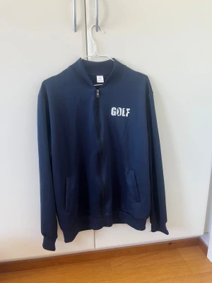 Mörkblå ziptröja GOLF tryck - Mörkblå ziptröja med vit GOLF-logga på bröstet och ryggen. Tröjan har ribbade muddar, två fickor fram och dragkedja hela vägen. Skön och avslappnad passform, perfekt till streetwear. Materialet känns som bomullsmix. Storlek L men känns mer som en M