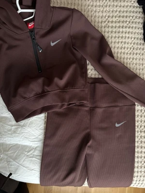 Brunt ribbat loungewear-set från Nike - Säljer ett brunt ribbat loungewear-set från Nike med croppad hoodie och matchande byxor. Hoodien har dragkedja framtill och Nike-logga på bröstet, byxorna har samma logga på benet. Oanvänd! 