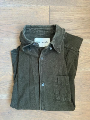 Mörkgrön skjorta från Our Legacy | Storlek 48 - Olivgrön skjorta från Our Legacy i bomull och linneblandning med diskret strukturmönster. Klassisk krage, bröstficka och knäppning framtill. Långärmad modell med normal passform, perfekt för en avslappnad men stilren look.