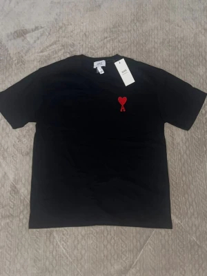 Svart t-shirt från Ami med rött hjärta - Snygg svart t-shirt från Ami med ett broderat rött hjärta på bröstet. Klassisk passform och rund halsringning. Perfekt för dig som gillar stilrena plagg med en twist. Materialet är mjuk bomull som känns skönt mot huden.