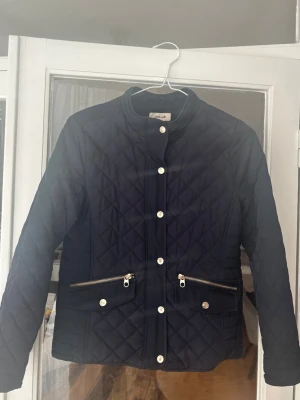 Mörkblå quiltad jacka med dragkedjor - Snygg marinblå quiltad jacka från Zara med guldfärgade knappar och dragkedjor på fickorna. Jackan har lång ärm, rund halsringning och ett stilrent rutmönster. Perfekt för dig som gillar klassisk men trendig stil. Jackan har använts ganska mycket. Knapparna är lite bortnötta i färgen. 