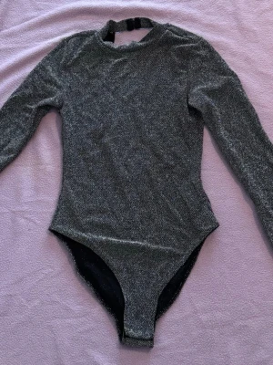 Glittrig svart långärmad body - Snygg svart body med glittereffekt och långa ärmar. Hög hals med två knappar bak i nacken. Stretchigt material som sitter tajt och ger en cool look. Perfekt för dig som vill sticka ut lite extra.