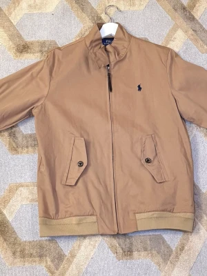 Beige jacka från Polo Ralph Lauren - Snygg beige jacka från Polo Ralph Lauren med blå broderad logga på bröstet. Jackan har dragkedja, två fickor med knappstängning och ribbade muddar. Insidan är fodrad med blått rutigt tyg. Perfekt för dig som vill ha en klassisk och stilren look.