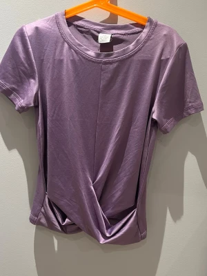 Lila omlottopp med twistad detalj - Säljer en lila topp med korta ärmar och snygg twistad omlottdetalj framtill. Toppen har rund halsringning och är gjord i mjukt, stretchigt material som känns skönt mot huden. Perfekt för dig som vill ha en enkel men trendig look.
