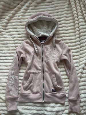 Rosa hoodie med dragkedja Superdry - En rosa hoodie med en dragkedja ifrån superdry i storlek M, passar mer som S enligt mig! Skönt och varmt material inuti! Defekter som mycket urtvättad, lösa trådar, en liten fläck och ett litet hål, alla syns på bild! Sen ser även det varma inuti ut o vara smutsigt, men det är sånt som fastnat i materialet.