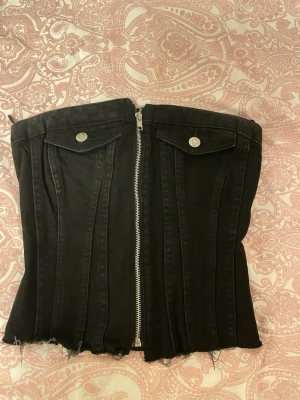 Zara jeans topp - Säljer en svart bustier i denimliknande material med silverfärgad dragkedja framtill och två dekorativa bröstfickor med knappar. Modellen är axelbandslös och har råa kanter nedtill för en edgy look.