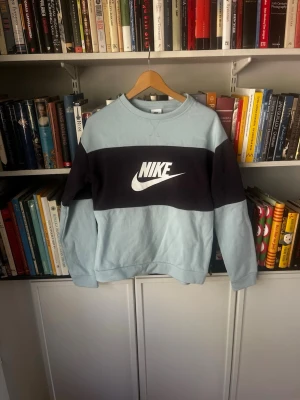 Vintage Nike sweatshirt i ljusblå och svart - Nike sweatshirt i ljusblått med bred svart panel över bröstet och stort vitt Nike-logotyptryck. Tröjan har rund halsringning, ribbade muddar och är tillverkad i mjuk bomullsblandning. Perfekt för en avslappnad streetwear-look.