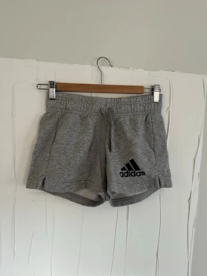 Grå shorts från Adidas med snörning - Grå mjuka shorts från Adidas med svart logga på vänster ben. Modellen har elastisk midja med snörning. Midja ca 33 cm och längd ca 31 cm bak. 