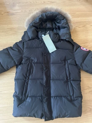 Svart dunjacka från Canada Goose med tag - Snygg svart dunjacka från Canada Goose med stor huva och äkta pälsdetalj. Jackan har raka linjer, två stora fickor framtill och klassisk logga på ärmen. Perfekt för kalla vinterdagar och riktigt varm tack vare dunfyllningen.sleek and functional cold-weather essential, this black puffer jacket combines style with serious warmth. It features a quilted, insulated design that provides excellent ej original heatretention while maintaining a lightweight feel. The jacket includes a high collar.