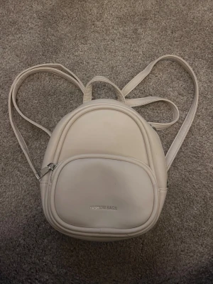 Beige miniryggsäck i skinnimitation - Säljer en stilren beige miniryggsäck från Fashion Bags i skinnimitation. Väskan har rundad form, justerbara axelremmar och ett stort fack samt en mindre ficka framtill med dragkedja. Perfekt för dig som vill ha en enkel och snygg accessoar.