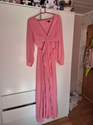 Lång rosa klänning med volangdetaljer - Säljer en elegant lång rosa klänning med v-ringning, markerad midja med dekorativ ring och volangdetaljer framtill. Klänningen har långa ärmar med resår vid ärmslut och ett mjukt, flytande tyg som ger en romantisk känsla.