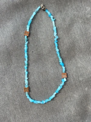 Turkost pärlhalsband med SNS-brickor - Turkost halsband med små oregelbundna pärlor och tre fyrkantiga SNÖ- brickor i metall. Halsbandet har ett klassiskt lås och pärlorna är av plast eller sten. Perfekt för dig som gillar färg och unika detaljer.