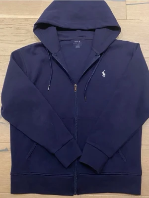 Mörkblå hoodie från Polo Ralph Lauren - Mörkblå hoodie med dragkedja från Polo Ralph Lauren. Klassisk design med huva och dragsnören, samt den ikoniska vita logon broderad på bröstet. Tillverkad i mjukt bomullsmaterial och har två fickor framtill. Perfekt för en avslappnad och stilren look. 