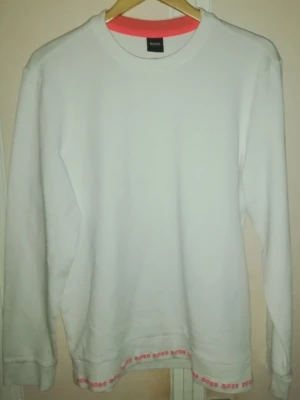 Hugo Boss sweatshirt vit - Vit sweatshirt från Hugo Boss med långa ärmar och rund halsringning. Tröjan har en diskret logotyp broderad framtill på bröstet. Längs nederkanten och insidan av halsringningen finns en kontrasterande detalj med upprepade BOSS-logotyper som ger plagget en stilren och unik design. Bekväm modell som passar både till vardag och casual stil.