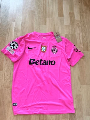 Sporting CP rosa matchtröja Nike - Sporting CP:s officiella rosa matchtröja från Nike med klubbmärke, Portugal-emblem och Betano-logga på bröstet. Tröjan har Champions League-märke på ärmen och är tillverkad i lätt, ventilerande polyester. Perfekt för fotbollsträning eller att bära på läktaren.