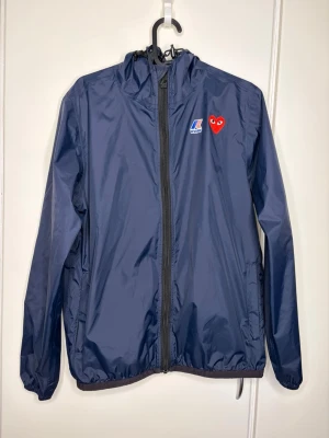 Blå vindjacka K-Way x Comme des Garçons - Snygg blå vindjacka från K-Way i samarbete med Comme des Garçons Play. Jackan har huva, hel dragkedja och två fickor. På bröstet finns K-Way-logga och det ikoniska röda hjärtat från Comme des Garçons. Perfekt för blåsiga dagar och streetwear-stil.