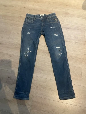 Blå slitna jeans från Replay - Säljer ett par blå jeans från Replay med skinny fit och slitna detaljer framtill STORLEK 30W 32L. Klassisk femficksmodell med dragkedja och knapp. Jeansen har en snygg tvätt och Replay-logga på bakfickan. Perfekta för en avslappnad och trendig look.