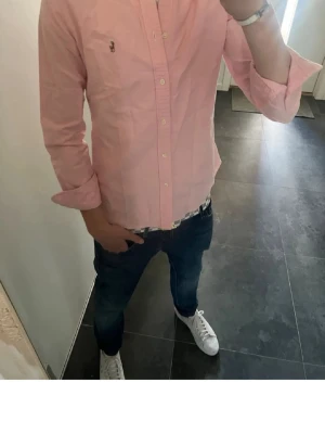 Rosa skjorta från Ralph Lauren - Snygg rosa skjorta från Ralph Lauren med klassisk krage och knappar framtill. Skjortan har lång ärm och den ikoniska broderade loggan på bröstet. Perfekt för en stilren och fräsch look. Passar bätter på s än m