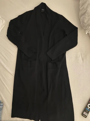 Svart lång cardiganjacka från Lindex - Svart lång cardiganjacka från Lindex med öppen front och två stora fickor framtill. Jackan har lång ärm och är tillverkad i ett mjukt, stickat material som ger en avslappnad och stilren look.