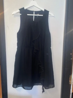 Svart ärmlös klänning med volang - Svart ärmlös klänning från H&M med volangdetalj framtill. Klänningen har en lätt och luftig känsla, markerad midja och rund halsringning. Perfekt för dig som gillar enkel men stilren design.