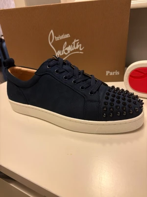 Blå Christian Louboutin sneakers med nitar - Mörkblå sneakers i mocka från Christian Louboutin med svarta nitar på tån och vit sula. Klassisk låg modell med blå skosnören och röd detalj under sulan. Perfekta för dig som vill sticka ut med en edgy och lyxig stil.