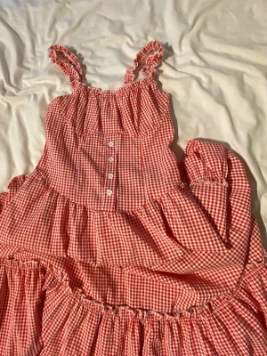 Rödvit rutig sommarklänning - Söt röd och vit rutig klänning med volanger och smockade axelband. Klänningen har knappar framtill och är sydd i ett mjukt, lätt bomullstyg. Perfekt för varma dagar och har en romantisk, lantlig känsla.