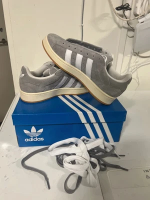 Grå Adidas sneakers med vita ränder - Snygga gråa Adidas sneakers i mocka med klassiska vita ränder på sidorna och vit sula med gummidetalj. Skorna har lågt skaft och kommer med både vita och grå skosnören. Perfekta för en stilren och sportig look.