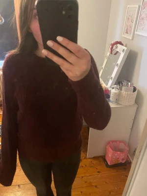 Vinröd fluffig stickad tröja - Mysig vinröd stickad tröja med rund hals och långa ärmar. Tröjan har en fluffig, mjuk struktur och ribbade muddar vid ärmslut och nederkant. Perfekt för kalla dagar och enkel att matcha med jeans eller kjol. Blivit något nopprig i materialet, använd fåtal gånger.