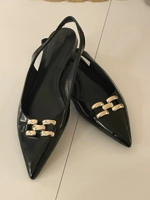 Svarta slingback skor ballerina stl 41 - Stilrena svarta ballerinaskor med spetsig tå och slingback-design. Skorna har glänsande guldfärgade metallspännen framtill som ger en lyxig touch. Materialet är blankt och elegant, perfekt för dig som gillar en chic look. 🖤🖤🖤Fint skick använda enstaka gånger. 