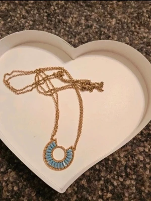 Guldhalsband med blå pärldetaljer - Unikt halsband med tunn guldfärgad kedja och ett runt hänge dekorerat med små blå pärlor i halvcirkelmönster. Hänget har en öppen cirkel i mitten och detaljerad guldram. Perfekt för dig som gillar färg och snygga accessoarer.