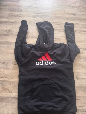 Svart Adidas hoodie med röd logga - Svart hoodie från Adidas med stor röd och vit logga på bröstet. Klassisk känguruficka framtill och huva. Perfekt för en sportig och avslappnad stil.