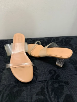 Klara sandaler med transparent klack - Snygga sandaler med två breda, transparenta remmar och fyrkantig tå. Klacken är i akryl och har en cool, genomskinlig look. Sulan är beige och ger en clean vibe. Perfekta för dig som gillar trendiga och minimalistiska skor.
