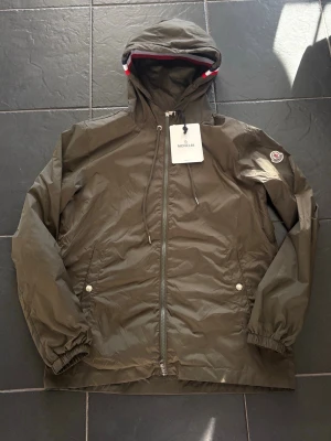 Olivgrön vindjacka från Moncler - Snygg olivgrön vindjacka från Moncler med huva och dragkedja framtill. Jackan har två fickor med tryckknappar, elastiska muddar och Moncler-logga på ärmen. Huva med röd, vit och blå randig detalj. Lätt och smidig i tunt material.