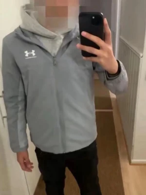 Grå vindjacka från Under Armour - Snygg grå vindjacka från Under Armour med dragkedja framtill och logga på bröstet. Jackan har en sportig look och är perfekt för dig som gillar en clean och enkel stil. Långärmad och med hög krage för extra komfort.