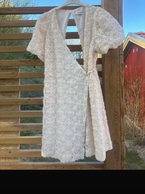 Studentklänning - Superfin cremevit kortklänning från Kappahl med 3D-blommor över hela tyget. Klänningen har omlottmodell, korta puffärmar och öppen rygg. Hade denna som studentklänning i fjol, så den är bara använd en gång. Nypris ca 700