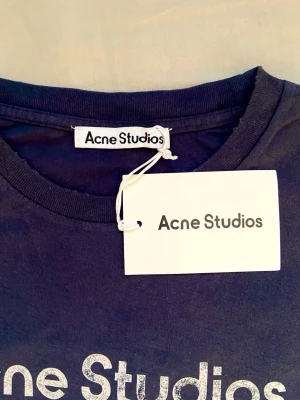 Blå långärmad topp Acne Studios - Snygg mörkblå långärmad topp från Acne Studios med tryck 'Acne Studios STOCKHOLM 1996' på bröstet. Klassisk rund halsringning och mjukt material, perfekt för en clean och stilren look.