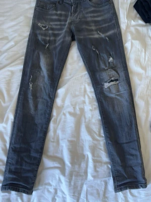 Grå slitna dsquared2 jeans - Säljer ett par grå dsquared2 jeans med slitningar och hål på benen för en trendig look. Jeansen har fem fickor, dragkedjegylf och är tillverkade i stretchigt denimtyg. Perfekta för dig som gillar en avslappnad stil.