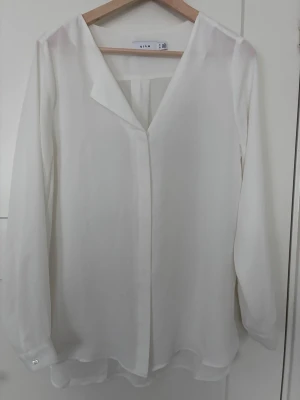 Vit v-ringad blus från Vila storlek M - Stilren vit blus från Vila med v-ringning och lång ärm. Tillverkad i ett lätt, transparent tyg som ger en luftig känsla. Blusen har en rak passform och knappar vid ärmsluten för en extra detalj. Perfekt att matcha med jeans eller kjol.